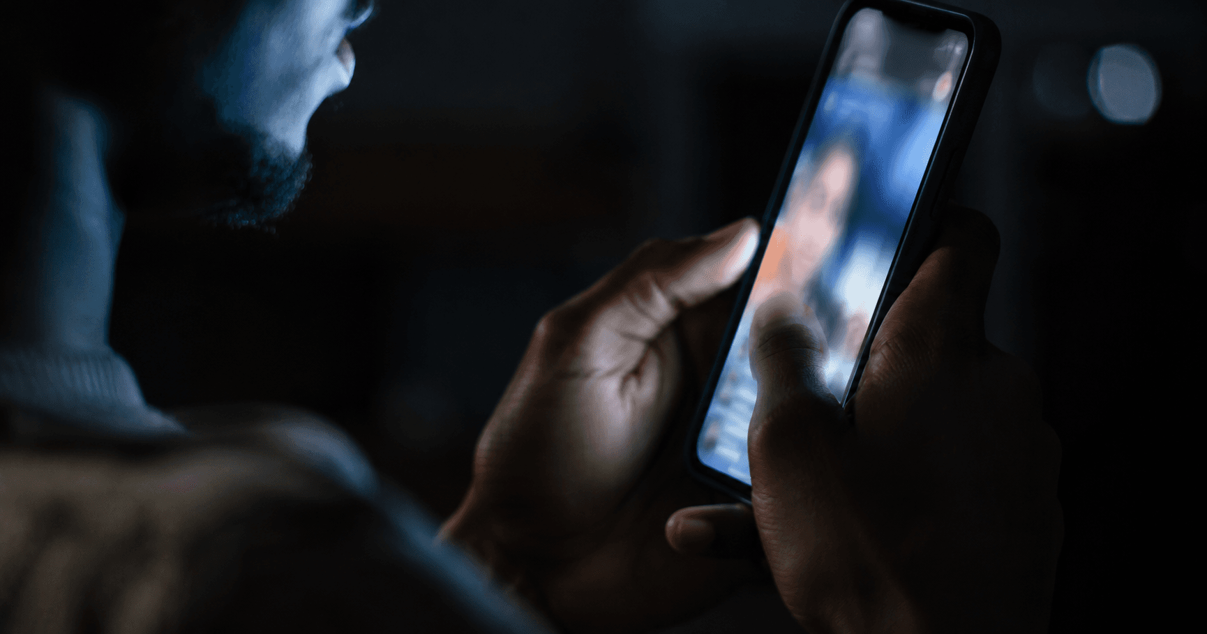 Hands scrolling a phone showing a blurred vertical video feed in dim lighting, representing short-form content consumption