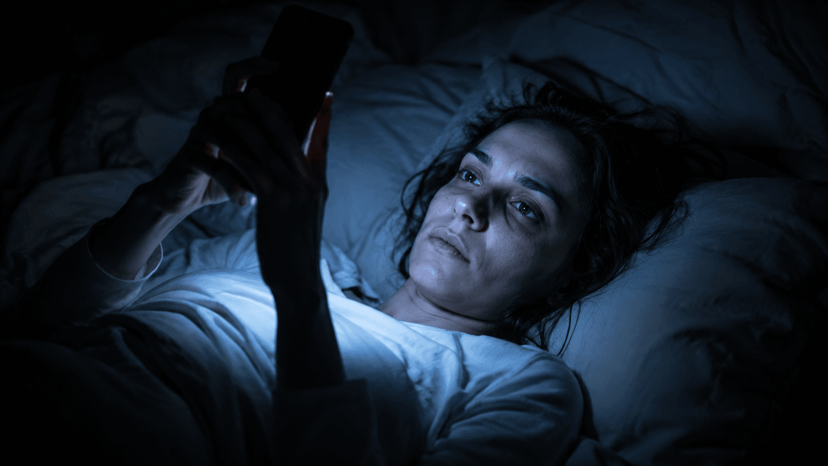 Person lying in bed at night, face lit by blue-white phone screen glow.